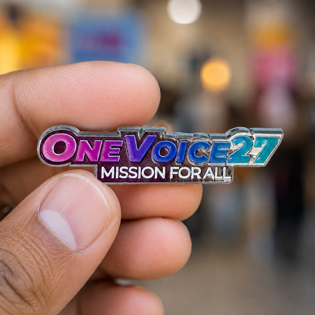 A close-up of a hand holding a silver enamel pin that reads 'ONE VOICE 2027' in pink, blue, and purple lettering, with 'MISSION FOR ALL' below it, against a blurred background.